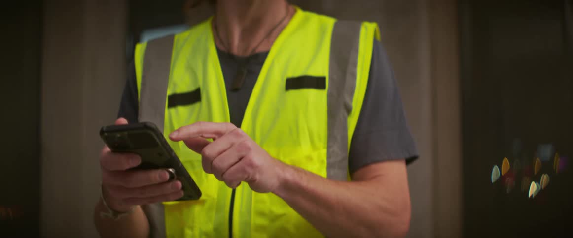 A man wearing a hardhat and safety vest stops tapping on his phone to wave his arm in instruction