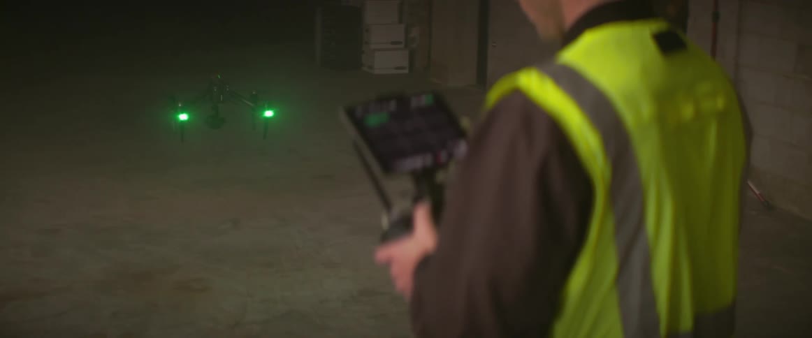 Worker controlling drone stands in building wearing a safety vest