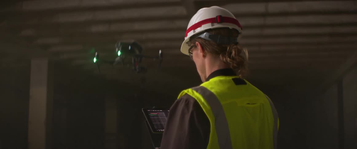A man stands inside a building and looks at a tablet as he controls a drone