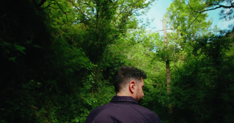 A man stands looking around at the trees scratching his head and rubbing the back of his neck