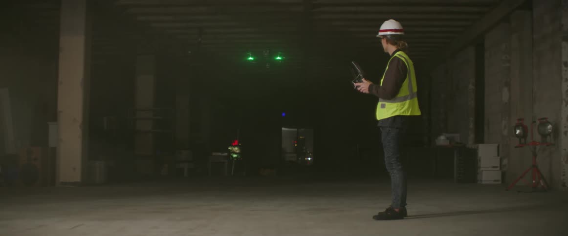 A man in a hardhat and safety vest controls a flying drone on a site