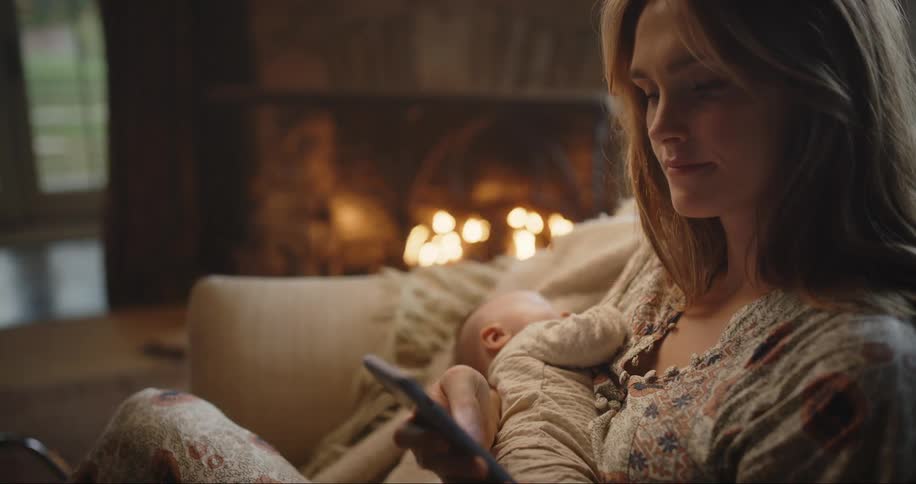 Mother sits holding baby in room with fireplace and scrolls on mobile phone