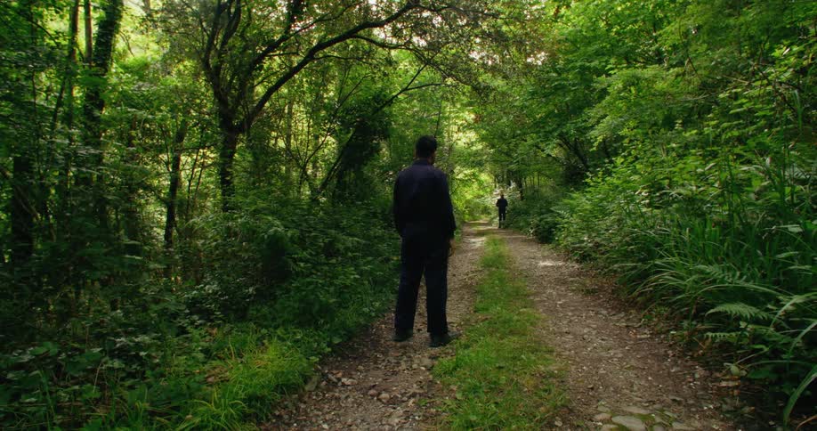Two men wearing black shirts and pants stand on dirt roadand staring at each other from distance in the woods