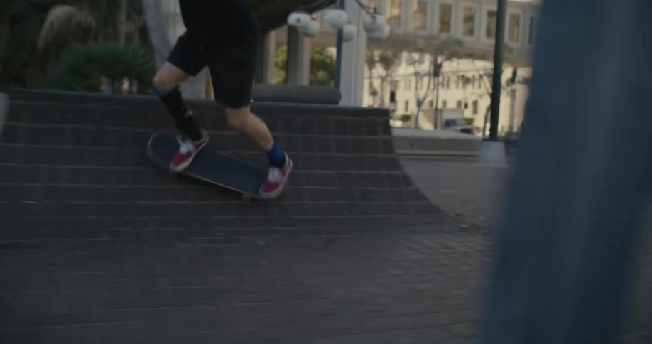 Skateboarder with a prosthetic brace fails to perform a trick as he rides near building with covered pedestrian bridge in the background