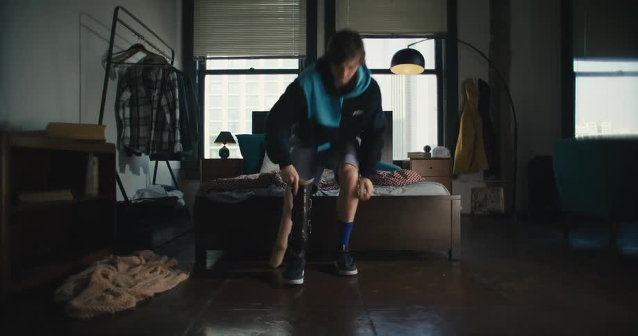 A man puts on his prosthetic leg while sitting at the foot of his bed in a studio apartment