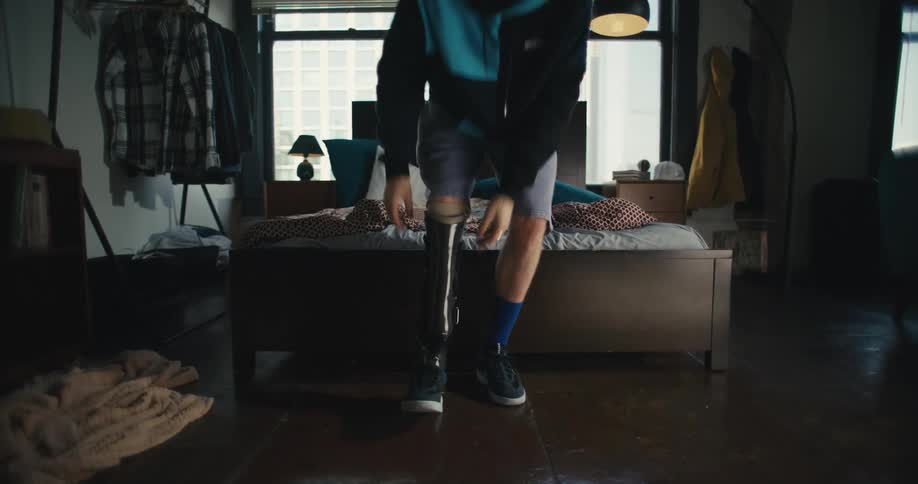 Man sitting on bed puts on prosthetic then walks away
