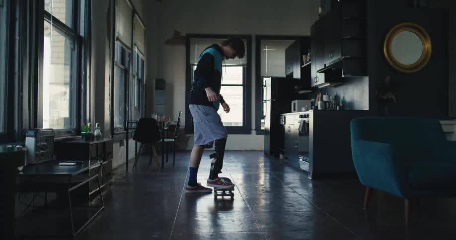 A young man with an artificial leg sets his skateboard down on his kitchen floor and falls when he stands on it and he then walks away