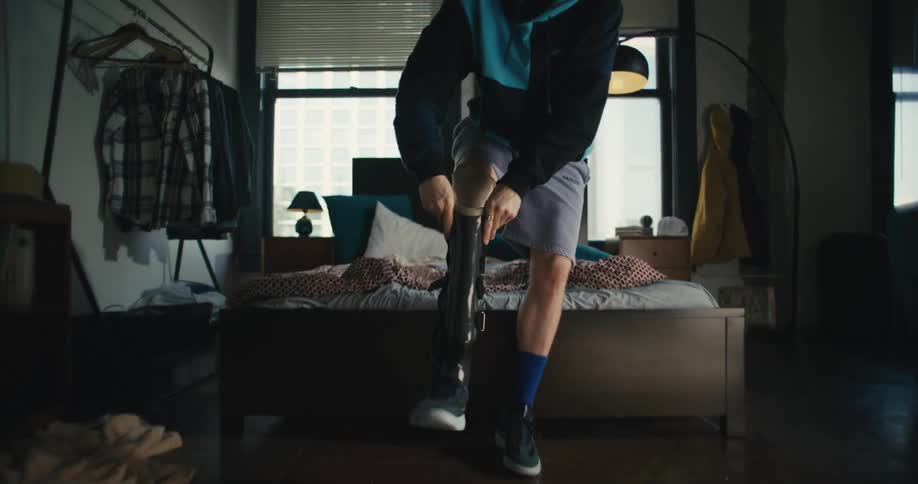 A young man sits on his bed and then stands up to put his artificial leg on