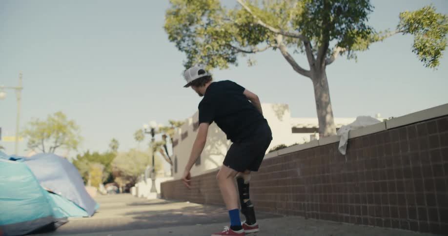 Male skateboarder wearing prosthetic almost falls while performing trick as he passes tents sitting on sidewalk
