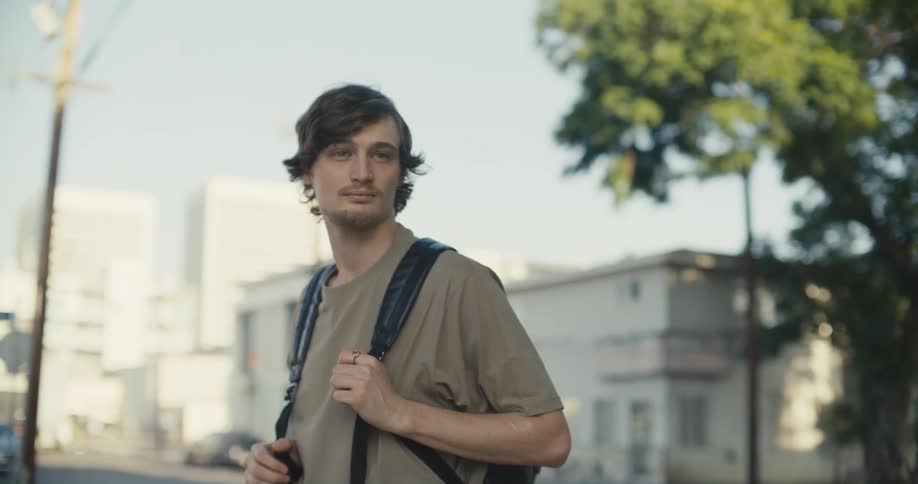 A teenager is standing outside holding the strap of his backpack and looking around as two people pass by him