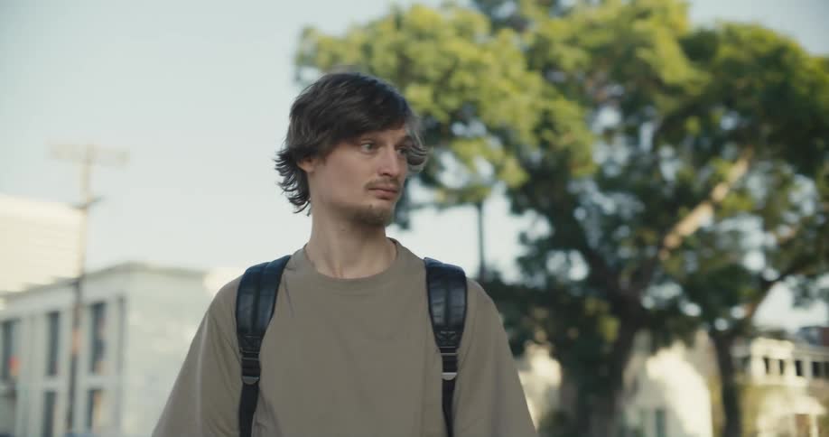 A teenager wearing a backpack is standing outside and looking around as two people pass by him