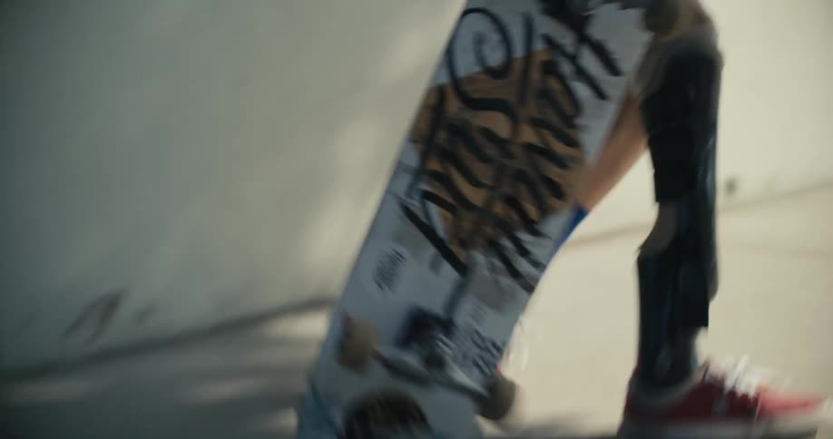A young man with an artificial leg sits on a skateboard on a sidewalk and rubs his face with his shirt then stands up and grabs his boards