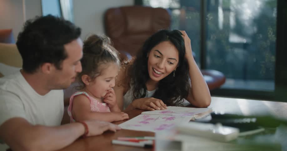 A mother and father sit at a table with their toddler daughter looking at a book