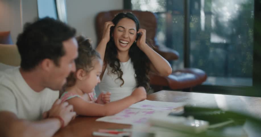 Toddler girl leans against coffee table looking at drawings in notebook between father with mustache white t shirt and mother with wavy hair and sleeveless white shirt