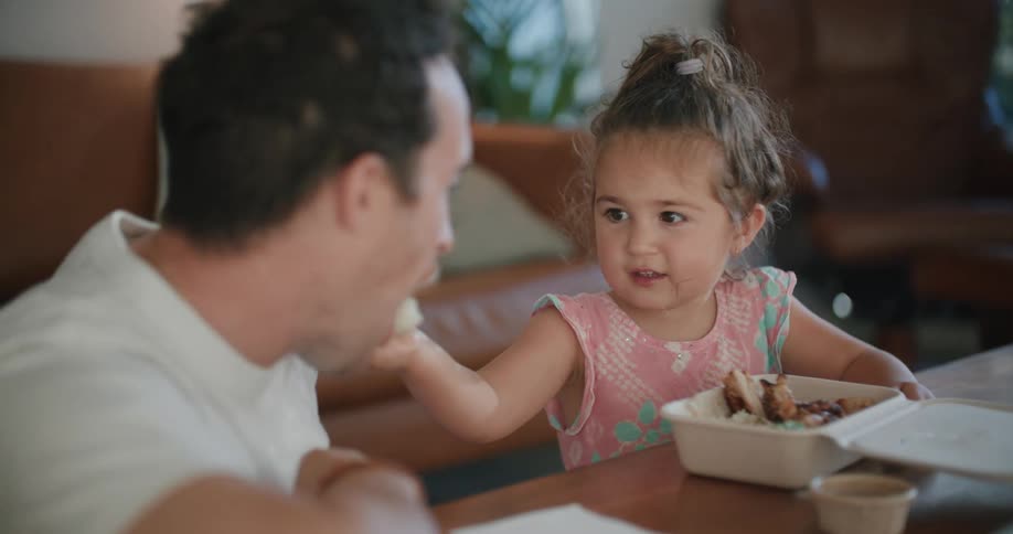 A man sits next to his young daughter at home and she gives him a spoonful of her food