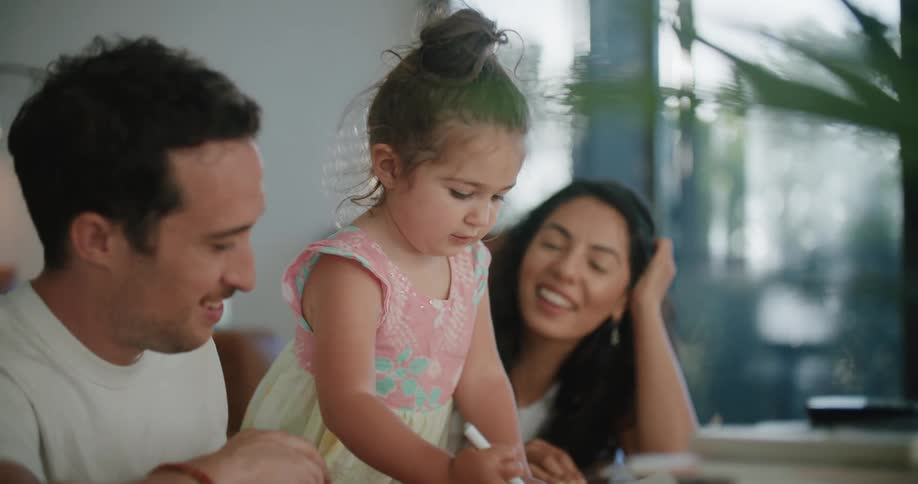 Father with mustache and white t shirt and mother with wavy hair look at daughter making a drawing in notebook in the living room at home