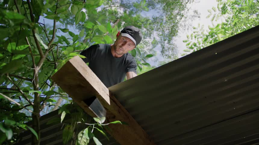 A carpenter saws wood off the end of a roof beam outdoors