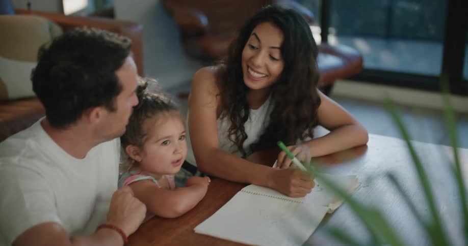 Woman with wavy hair white sleeveless shirt and jeans writes in notebook sitting at coffee table with father and toddler girl in living room