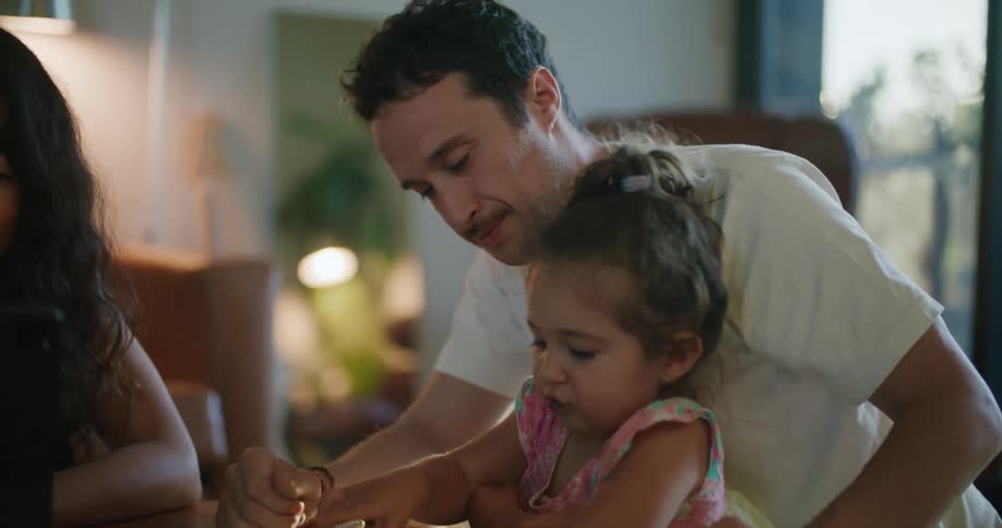 Father with mustache white t shirt holding toddler girl making a drawing in notebook next to mother with wavy hair using tablet at coffee table