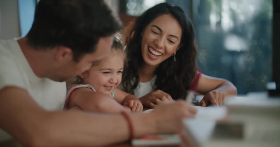 Toddler girl leans against coffee table watching father with mustache white t shirt making a drawing in notebook next to mother with wavy hair and sleeveless white shirt