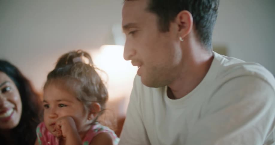 Toddler girl leaning against the coffee table watches tablet between father with mustache white t shirt and mother with wavy hair and sleeveless white shirt talking with each other