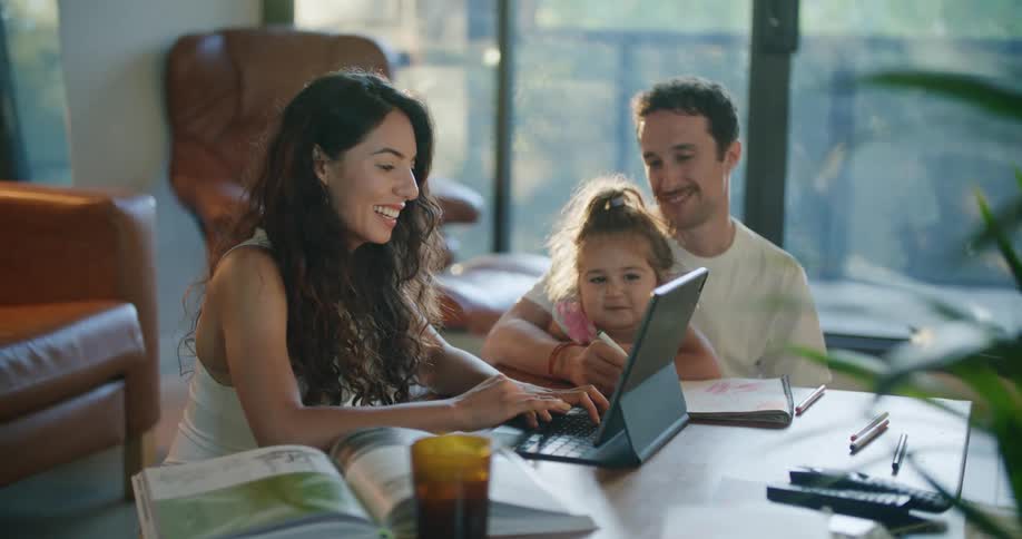 Father with mustache and white shirt holds daughter making a drawing in notebook sitting at coffee table with mom with wavy hair and sleeveless shirt using tablet in living room at home