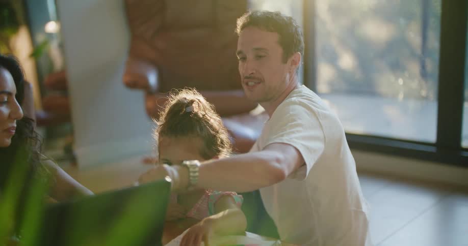 Father with mustache white t shirt holding toddler girl making a drawing in notebook next to mother with wavy hair using tablet at coffee table