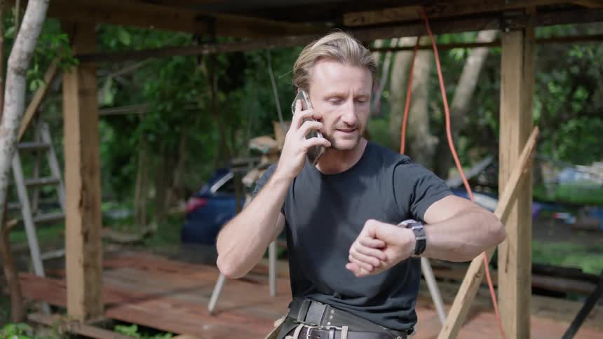 A man is sitting down outside at a work site and he makes a phone call