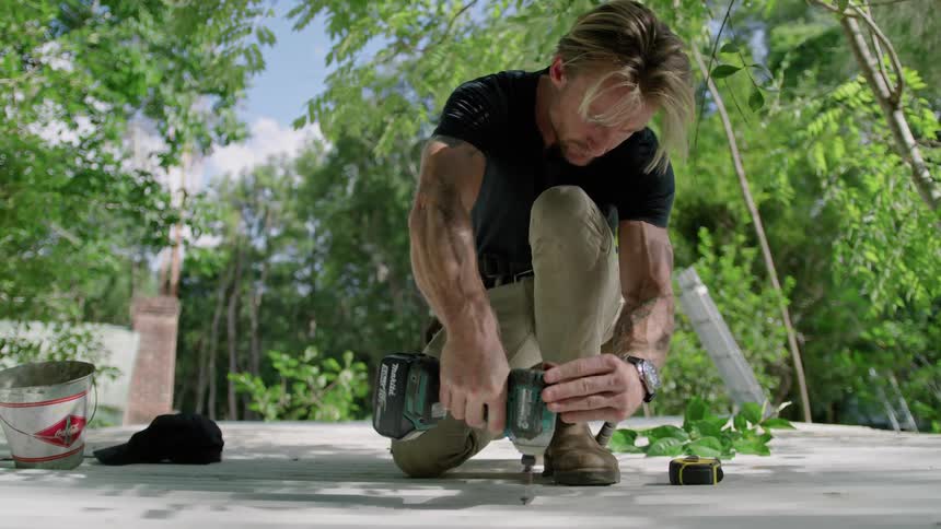 A man is working outside using a hand drill and there is a forest of trees around him