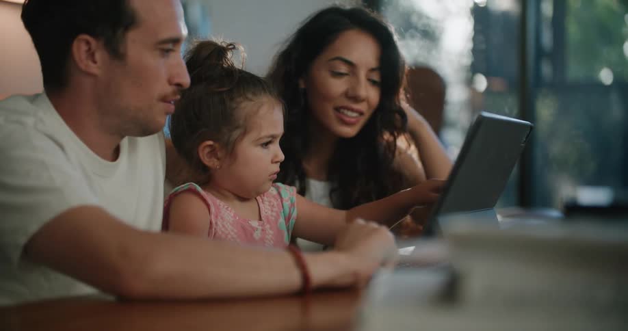 A mother and father sit with their toddler daughter in front of a tablet