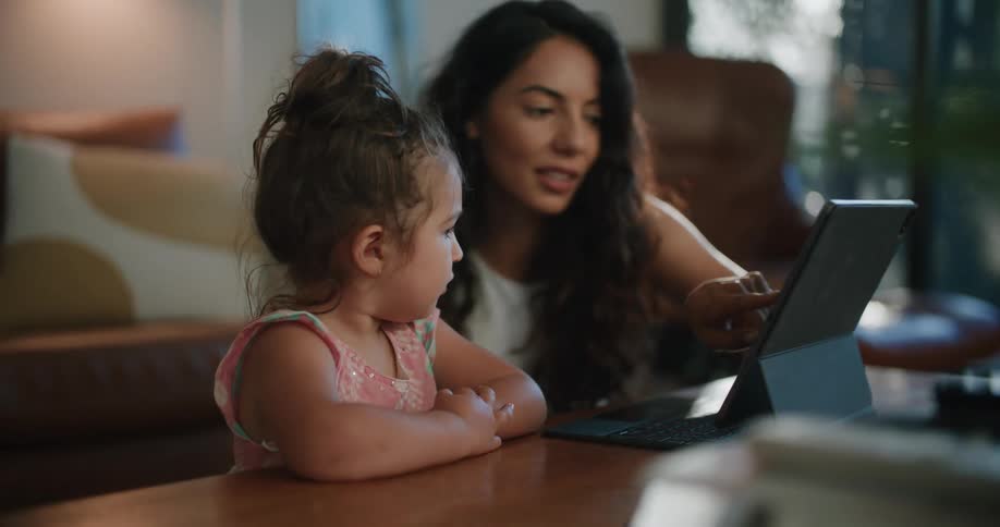 Mother with wavy hair and white sleeveless shirt uses tablet next to daughter leaning against coffee table in living room at home