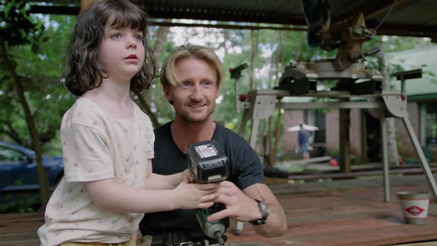 A father hands a drill to his daughter to carry towards someone