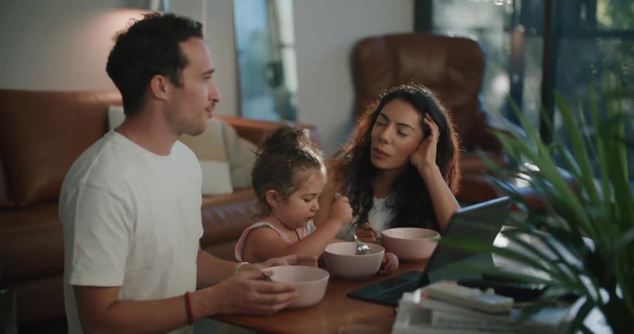A man and woman sit in their living room eating with their young daughter at a table with a laptop on it