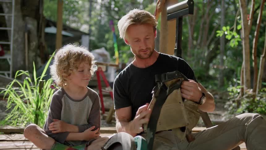 A man shows his young son tools from a tool belt as they sit outside
