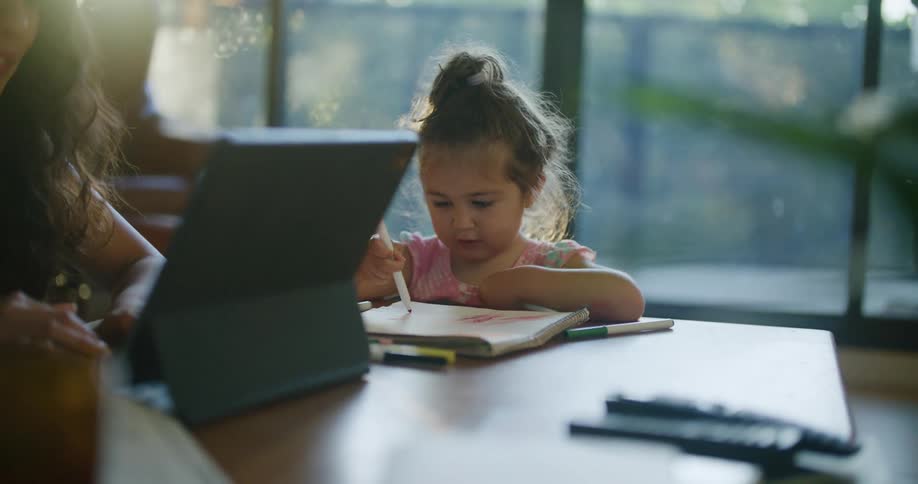 Mother with wavy hair and sleeveless white shirt uses tablet next to daughter making a drawing in notebook in living room at home