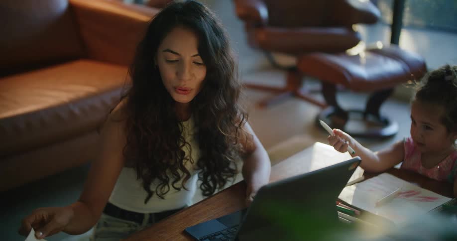 Mother with wavy hair and sleeveless white shirt uses tablet next to daughter making a drawing in notebook in living room at home