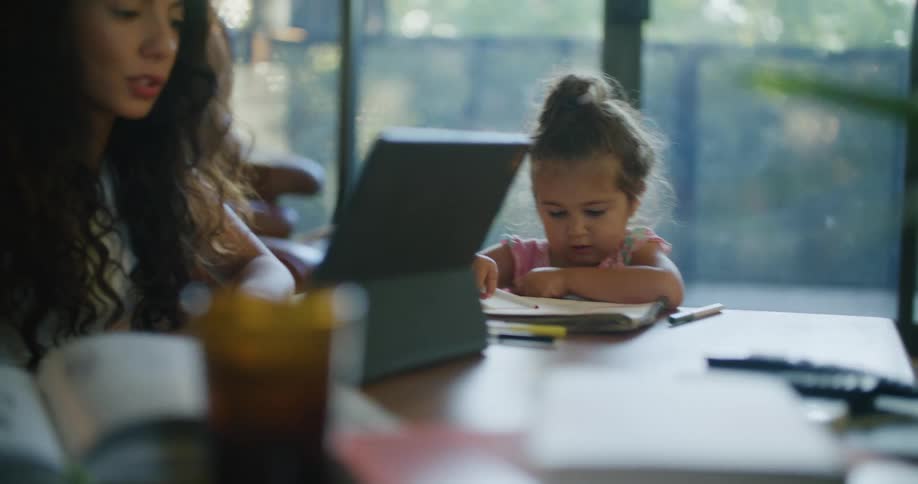 Woman working on laptop while daughter coloring next to her on kitchen table