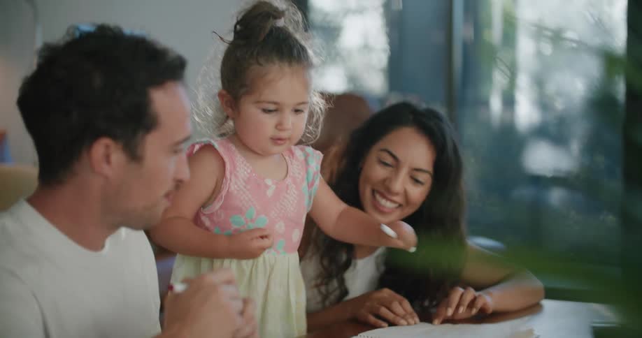Father with mustache white t shirt toddler girl and mother with wavy hairand white sleeveless shirt make drawings in notebook at coffee table in living room at home