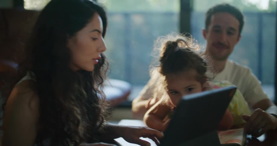 Father with mustache holds daughter watching mother with wavy hair using tablet at coffee table in living room at home