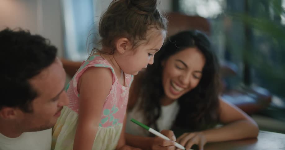 Mother with wavy hair sleeveless white shirt makes a drawing in notebook next to toddler girl and father with mustache and white t shirt in living room at home