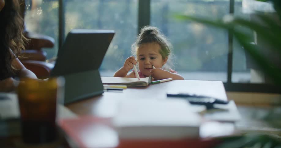 A woman holds a video call on her laptop while her daughter draws nearby