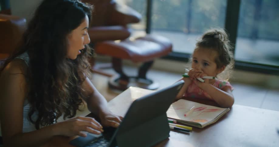 A toddler draws while seating next to her mother on a laptop