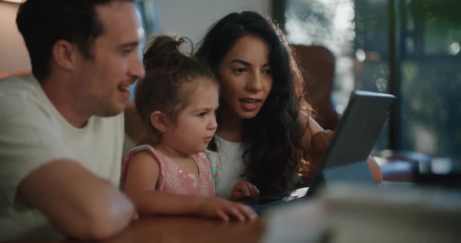 Father with mustache and white t shirt and mother with wavy hair and sleeveless white t shirt help daughter use tablet at coffee table in living room at home