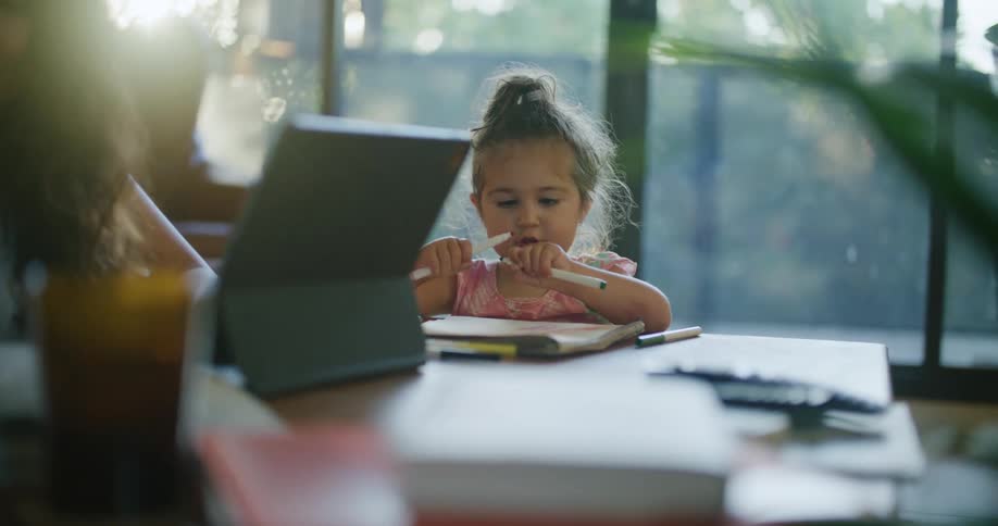 A woman reads a magazine and uses her laptop while her daughter draws beside her
