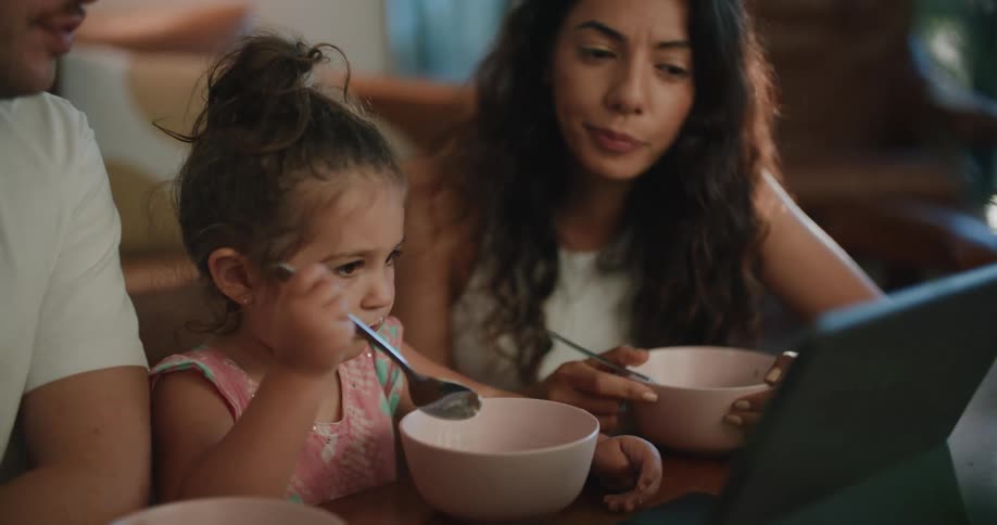 Mother with wavy hair white shirt toddler girl and father with white t shirt and mustache eat food out of bowls watching tablet at coffee table in living room