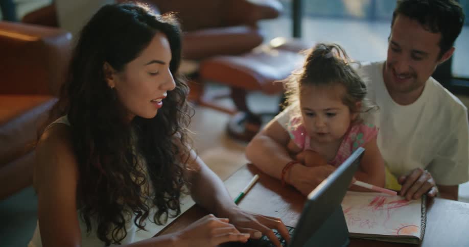 Father with mustache white t shirt holding toddler girl making a drawing in notebook next to mother with wavy hair using tablet at coffee table