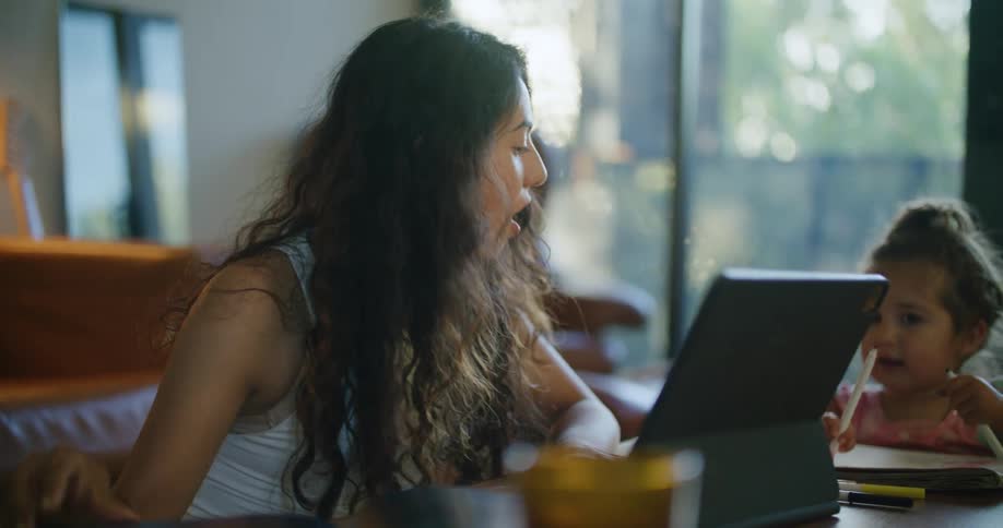 Mother with wavy hair and sleevelesss white shirt uses tablet sitting at coffee table next to daughter making a drawing in notebook in living room