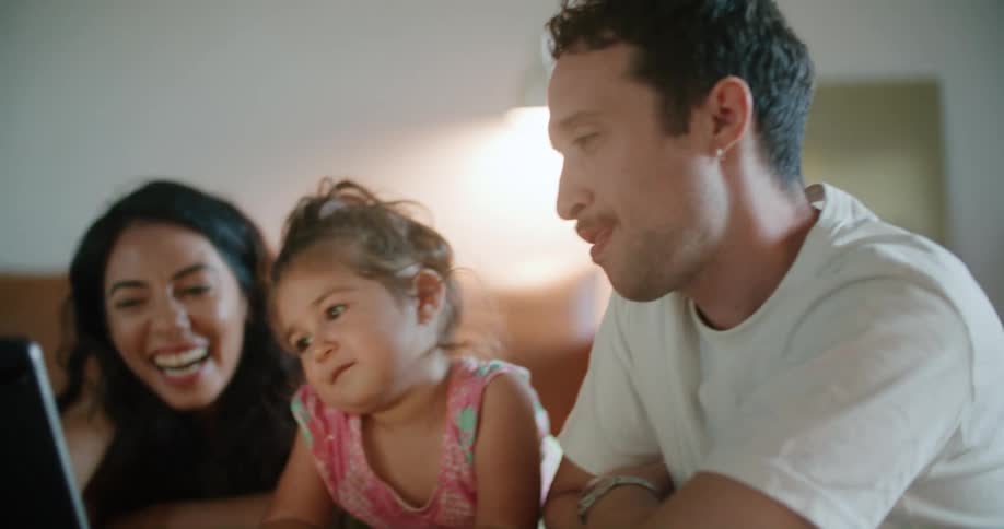 Toddler girl leaning against the coffee table watches tablet between father with mustache white t shirt and mother with wavy hair and sleeveless white shirt