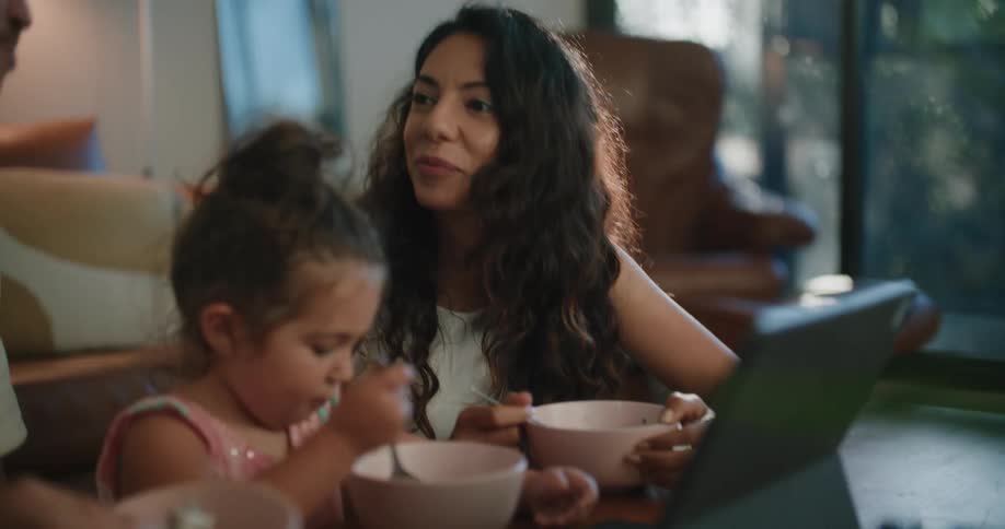 A family laughs while eating at their coffee table while their daughter watches a tablet