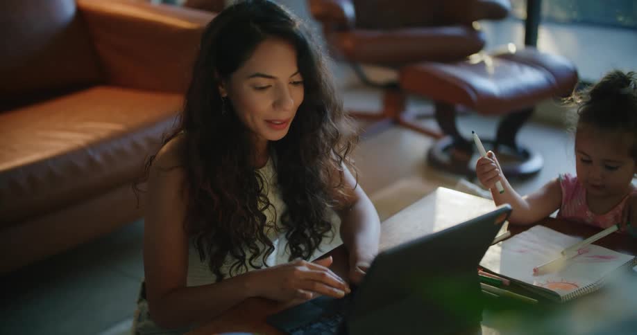 Mother uses laptop while smiling and talking with daughter who is holding markers